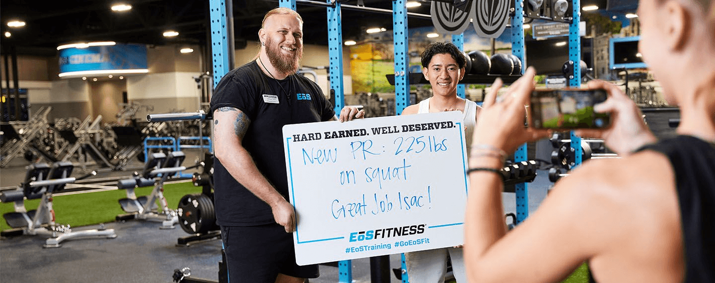 A gym member holding a whiteboard showing their new personal record while another person takes a photo with a smartphone