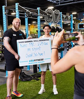 eos gym member and personal trainer getting their photo taken holding up how much weight they lifted