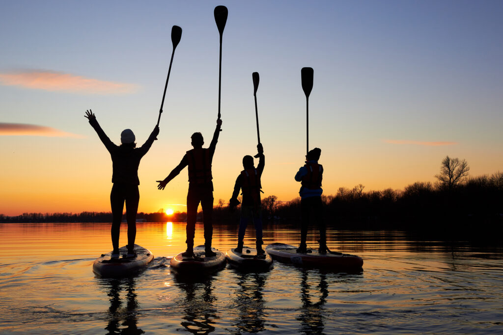 Four friends on stand up paddle board (SUP) on a flat quiet winter river at sunset raising their paddles up